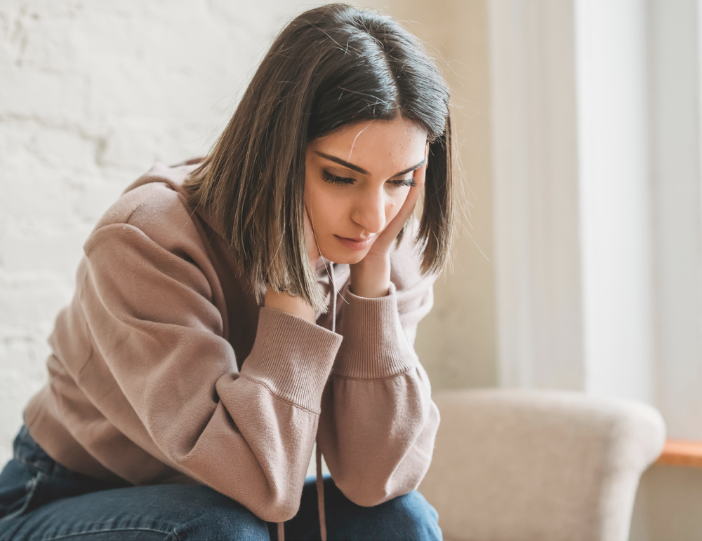 A young woman sitting with her head in her hands, looking tired, overwhelmed, and deep in thought. She appears physically and emotionally drained, capturing the feeling of chronic stress and burnout.