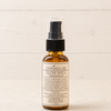A small amber glass spray bottle of Lifeholistically Glow Well Facial Serum with a black pump and clear cap, standing on a light wood surface against a light wood plank background.