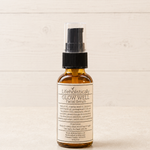 A small amber glass spray bottle of Lifeholistically Glow Well Facial Serum with a black pump and clear cap, standing on a light wood surface against a light wood plank background.
