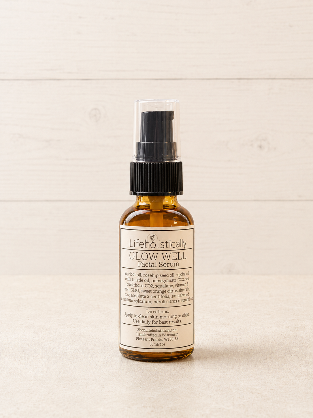 A small amber glass spray bottle of Lifeholistically Glow Well Facial Serum with a black pump and clear cap, standing on a light wood surface against a light wood plank background.